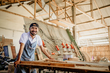 Naklejka na ściany i meble Asian entrepreneur in a hat smiling at the camera standing in a woodcraft workshop