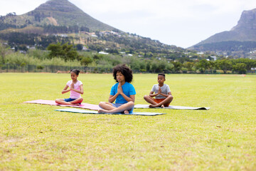 Happy african american schoolchildren doing yoga and meditating on field at school