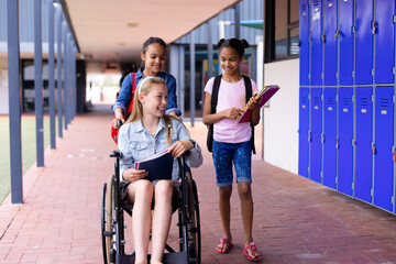 Happy diverse schoolgirl in wheelchair with her friends in corridor at school