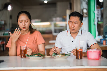 Asian woman and man sitting with a spicy expression while eating pecel in a traditional food stall