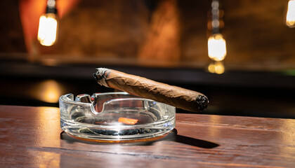 Burning handmade luxury Cuban cigar resting on an ashtray on an old wooden countertop in a...