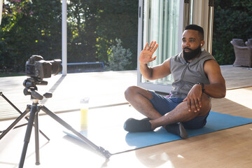 Happy african american man wearing sportswear on mat and making video blog, waving hand at home