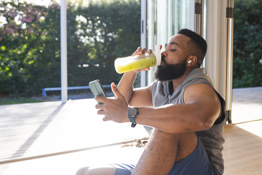 Focused african american man wearing sportswear, drinking juice and using smartphone at home