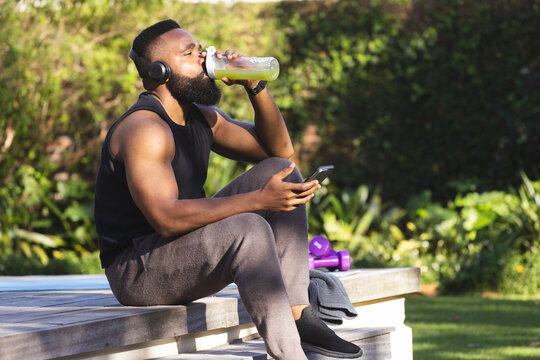 African american man wearing sportswear, headphones, drinking juice and using smartphone in garden