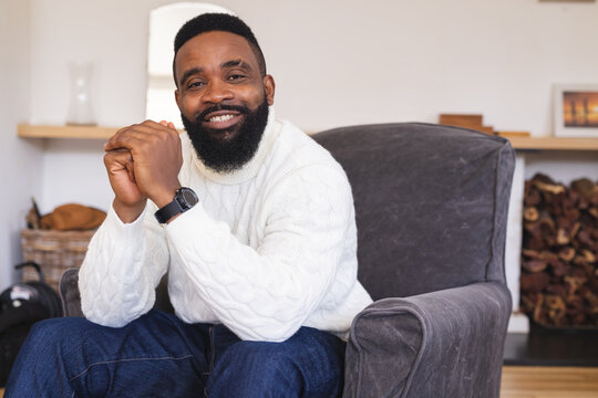 Portrait of happy african american man wearing watch, white jumper and sitting on armchair at home