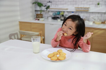 Kid asian black hair girl sitting on table and drinking milk while eating vanilla cookies for breakfast with enjoying time. Tasty food and delicious food with happy meal lifestyle kid concept