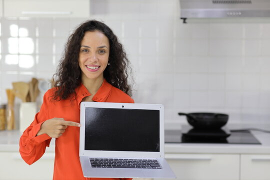 Smart and active latin woman standing while whowing and pointing at computer with happy smiling face in kitchen. Anywhere and work ifestyle with technology concept.