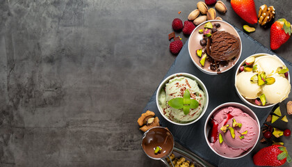 Fototapeta na wymiar Tubs of fresh fruit with ice cream and ingredients including, chocolate, berries, walnuts, pistachio and a metal scoop for serving on slate with copy space, top down view