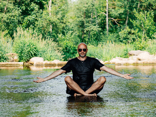 Bald male in dark wet shirt and shorts sitting in a stream and practice yoga relaxation. Selective focus. Green nature background. Healthy habit to battle stress and reach inner peace. Yoga guru.