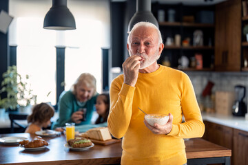 Satisfied happy senior man looking at camera and enjoying delicious breakfast, gesturing with hands standing in front of family members at kitchen table, senior woman and two kids at home.