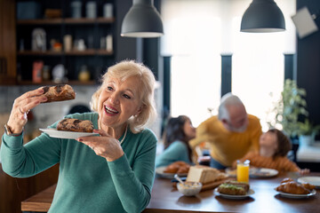 Beautiful happy senior woman in selective focus holding a plate and looking at sweet delicious pastries while standing in the kitchen with senior man and two kids playing at table in the background.