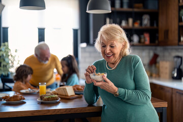 Smiling senior woman eating porridge for breakfast standing in the kitchen in selective focus in front of kitchen table with senior man talking to two small children in the background.