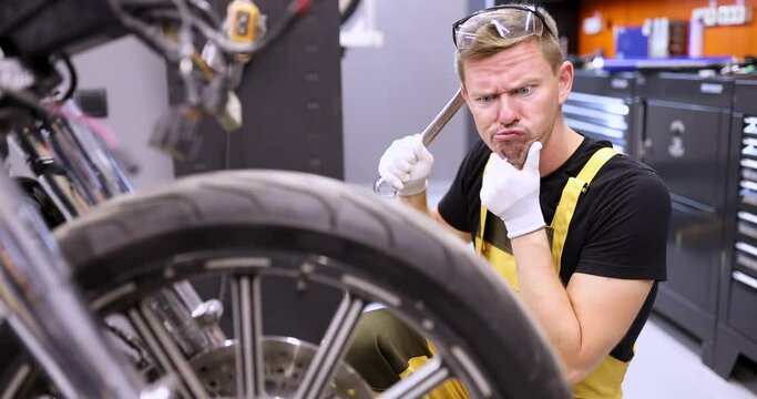 Emotionally pensive auto mechanic holds wrench and stares in surprise at motorcycle in service center. Auto transport service concept