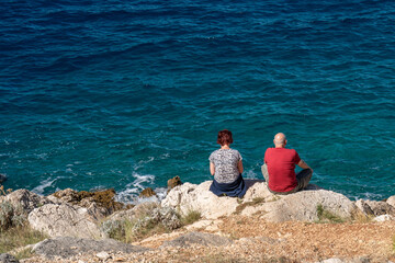 .A man and a woman sit on a rocky seashore and enjoy the beautiful view of the blue, transparent...