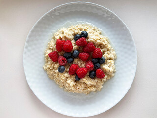 Ripe dark blue blueberries and red raspberries lie in a heap in a texture gray round plate on top of boiled oatmeal, close-up, top view. Plate with food on a gray surface. Proper nutrition. Vitamins.