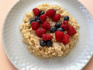 Ripe dark blue blueberries and red raspberries lie in a heap in a texture gray round plate on top of boiled oatmeal, close-up, top view. Plate with food on a peach surface. Proper nutrition. Vitamins.