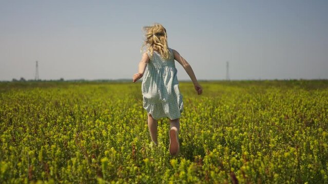Young girl in white dress runs through yellow wildflower field, following from behind