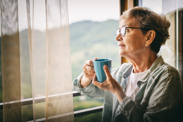 Beautiful senior elderly woman standing at the window overlooking the mountains and holding a mug...