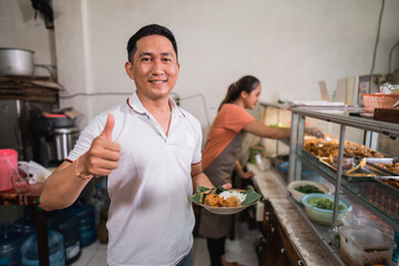 handsome male customer carrying a plate of pecel food with thumbs up standing at a traditional stall
