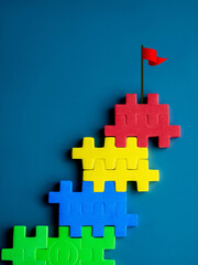The ultimate goal, red flag on top of colorful puzzle blocks as bar graph chart steps on blue...