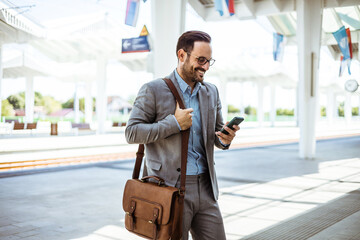 Photo of a smiling businessman texting on his mobile phone at train station. Businessman on commute transit talking on the smartphone while walking with hand luggage in train station