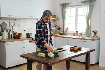 A standing elderly Asian man examining the ingredients on the table while scrolling through cooking videos on his smartphone in the kitchen.