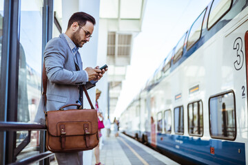 Handsome young man on business trip walking with his luggage and talking on cellphone at train station. Travelling businessman making phone call. Photo of a smiling businessman at train station - obrazy, fototapety, plakaty