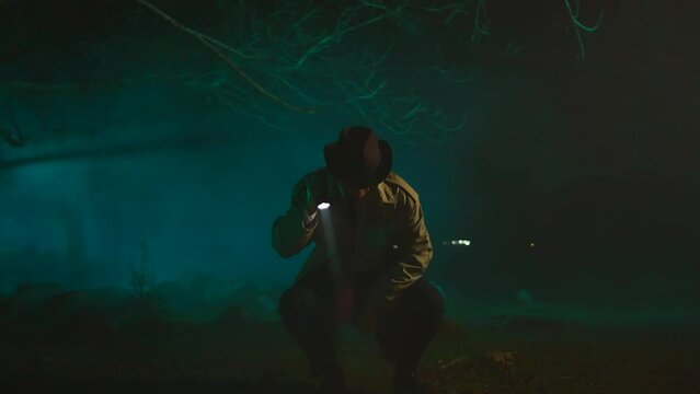 Old fashioned detective a man in a hat with a flashlight in his hands in a dark forest is searching for clues