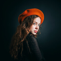 Portrait of a sad young woman in an orange beret and long curly hair on a dark background. - 622579244