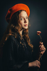 Portrait of a sad young woman in an orange beret with a flower in her hand on a dark background. - 622579423