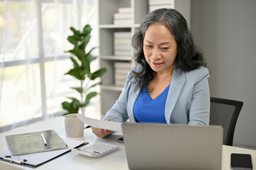 A happy Asian senior female boss reading business documents and working on her tasks
