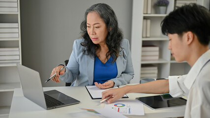 A serious Asian senior businesswoman focuses on working with a young male worker