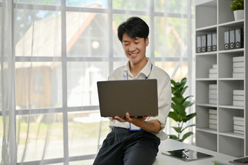 A handsome young Asian male office worker or banker sits on a table and uses his laptop