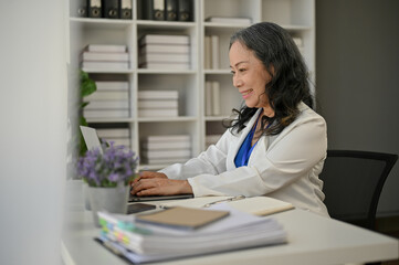 Obraz na płótnie Canvas A happy Asian senior businesswoman working on her business tasks on her laptop at her desk
