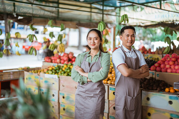 a couple of fruit seller with apron standing together with handcrossed with the fruits at the background