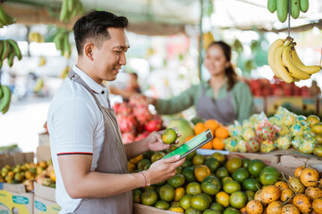 male fruit seller looking at the digital tablet while checking the oranges with the female seller working at the background