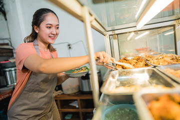 female stall owner wearing apron using food tongs to pick up side dishes at a food stall display