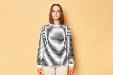 Serious strict brown haired woman wearing striped shirt standing isolated over beige background...