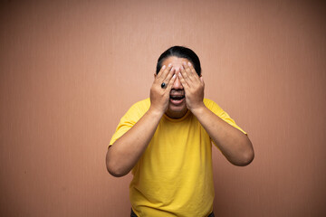 old asian man wearing yellow tshirt covering his face because feeling afraid or fear in plain...