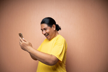 old asian man wearing yellow tshirt holding handphone and using it to texting or video call with...