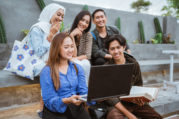 a group of Asian students look at a laptop while studying together with a laptop outside the campus