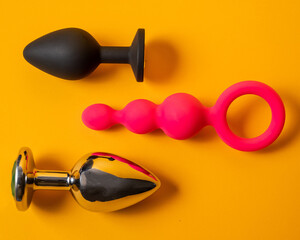 A set of three alternative sex devices of various shapes and sizes on an orange background. Silicone and metal plugs and anal beads. 