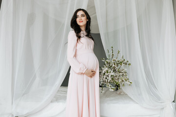 A beautiful pregnant woman in a pink dress holds her hands on her stomach. A loving mother is...