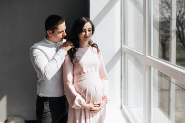 A pregnant woman and a man hug by the window. Support of a husband to a pregnant wife