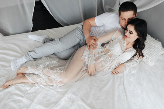 Young brunette pregnant woman in her room in a white lace dress on the bed with her husband. Motherhood. Concept of pregnancy, motherhood, preparation and waiting.