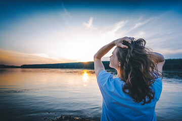 Young relaxed woman with wavy hair standing with her back on the river bank at sunset. Holding my...