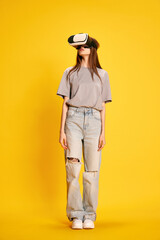 Full-length portrait of young beautiful girl in casual clothes, wearing vr glasses against yellow studio background. Concept of human emotions, fashion, youth, lifestyle, female beauty, ad