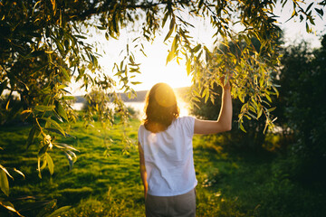 Young woman standing with her back in the rays of the sun at sunset on the banks of the river at...