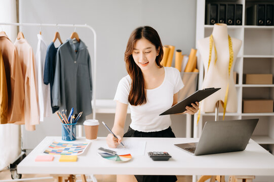 Asian young woman on desk in office of fashion designer and holds tablet, laptop and smartphone on white table