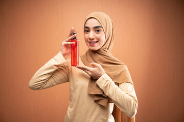 muslim woman wearing hijab and cream t-shirt holding skincare bottle smiling at camera in orange...